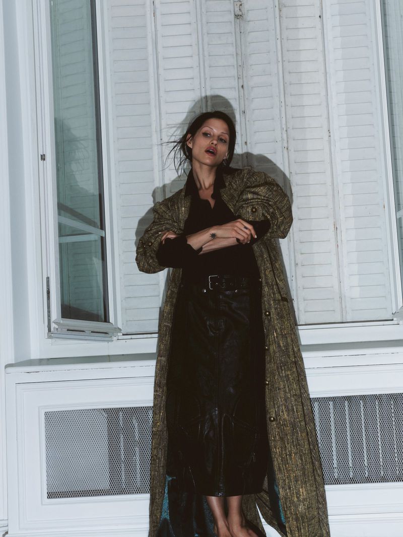 A woman in a long coat stands indoors near white shuttered windows, creating a fashion editorial shot with a neutral color palette.