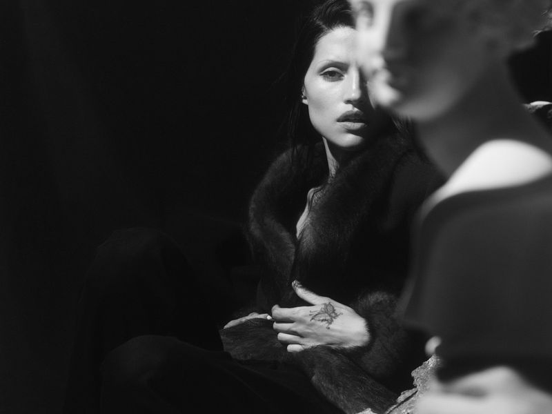 Monochrome portrait of a woman in a fur coat with a hand tattoo, partially obscured by a classical bust in soft studio light.