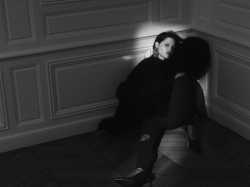 Black and white portrait of a woman in dark clothing, sitting in a corner with light catching her face, against a paneled wall.