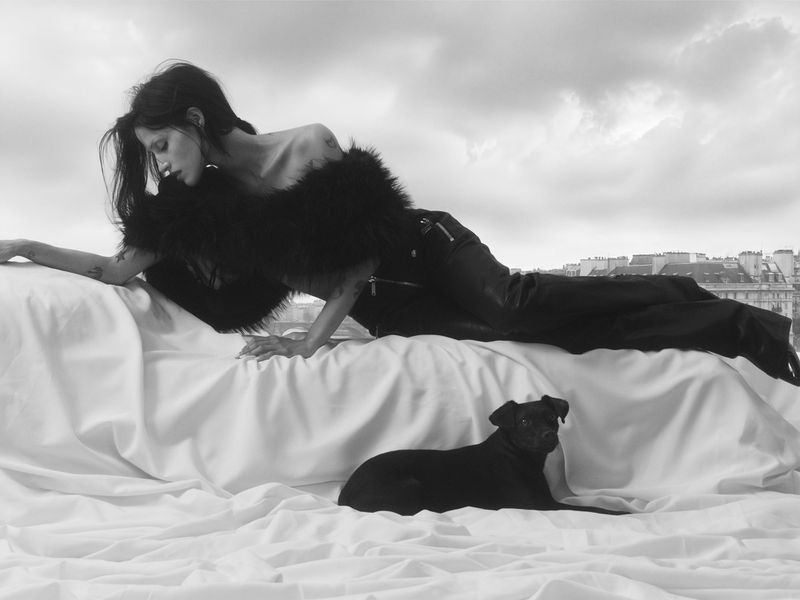 Monochrome portrait of a woman lying on a bed with a black dog, cityscape visible through the window in the background.