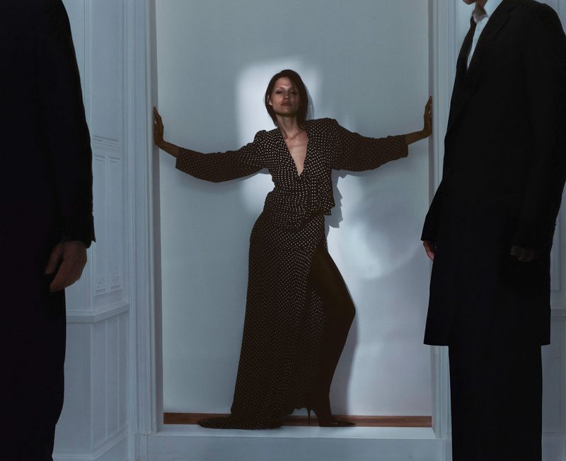 Stylish studio shot of a woman in a polka dot dress, posed between two men in suits against a white wall. Fashion editorial.