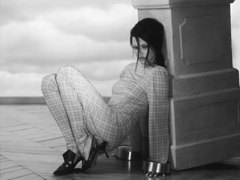 Monochrome portrait of a female in a plaid suit crouching with metal ankle restraints, leaning against a pillar against a cloudy sky.