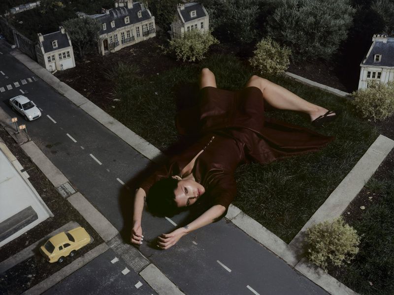 Woman in dress lies on grass in miniature town with model houses and cars, creating a surreal, oversized effect in an outdoor setting.