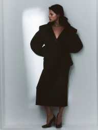 Model Gabbriette poses in an oversized black tailored blazer, midi skirt, and black high heels against a white studio wall.
