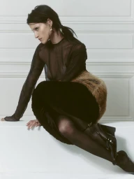 Model Gabbriette crouching on a white ledge wearing a sheer black top, faux fur accents, tights, and black patent boots.