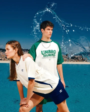Two models wearing vintage-inspired Umbro apparel with a water splash effect and cityscape backdrop on a bright, sunny day.