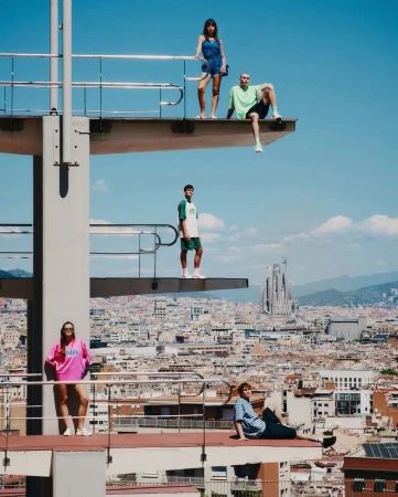 Five people in casual clothes stand on different levels of a high diving platform overlooking Barcelona cityscape with La Sagrada Familia.