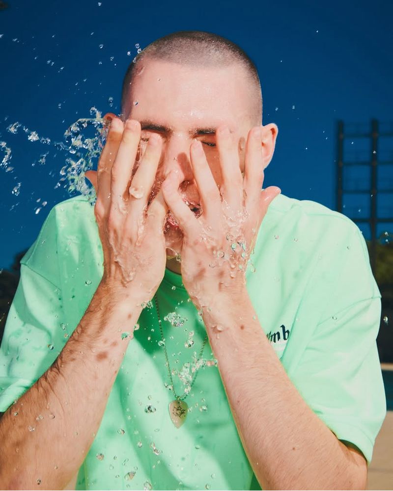 Close-up of a young adult man with his face covered by his hands as water splashes against his skin, with a blue sky background.