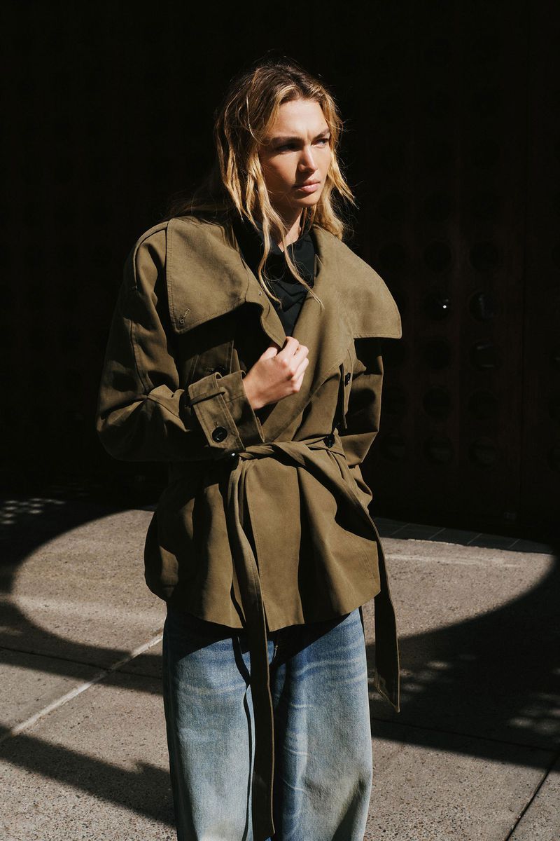 A blonde woman in a belted olive trench coat and wide-leg jeans stands in a shadowed outdoor setting on a sunlit paved surface.