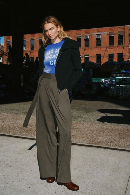 A young woman poses in a blue graphic tee, black zip jacket, and olive wide-leg pants on an outdoor urban catwalk with brick buildings.
