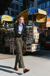 Model standing outdoors near a food cart wearing a black zip cardigan, blue t-shirt, and olive trousers