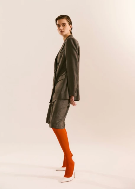 A young woman in a grey skirt suit poses in a studio wearing bright orange tights and white high heels for a fashion editorial.