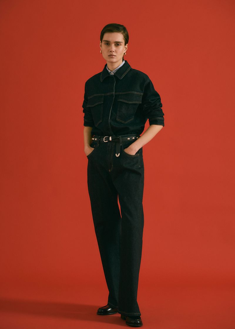 A woman poses in a dark-wash denim jacket and pants with a belt against a red background in a studio shot, showcasing a fashion editorial.