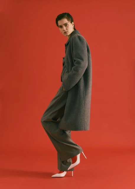 Fashionable woman poses confidently in coat and wide-leg pants on red backdrop. High heels add a touch of sophistication to the monochrome look.