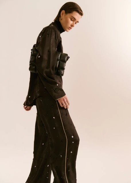 Androgynous person in a dark, embellished pajama suit, side angle in a studio, showcasing fashion and style.