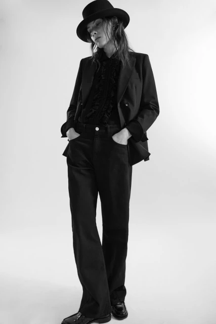 Black and white full shot of an adult woman wearing a hat, blazer, blouse, and pants against a white backdrop in a photo studio.