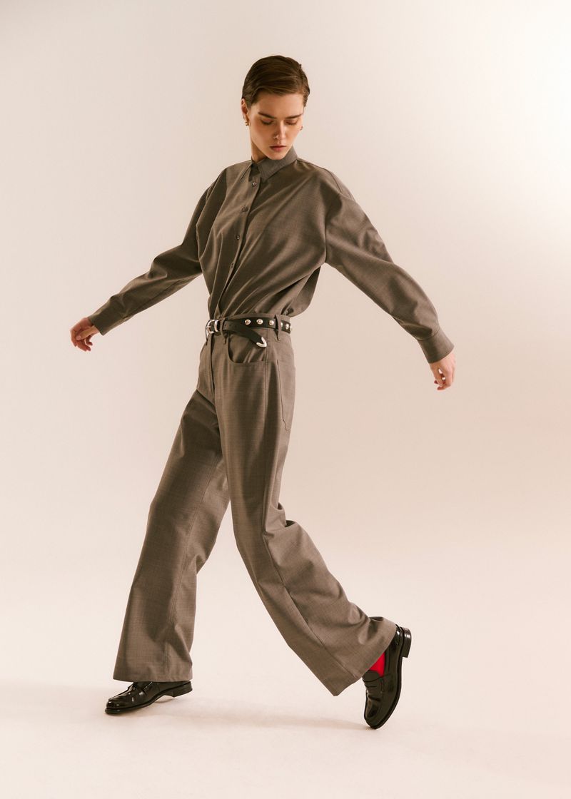 A young woman in a grey pantsuit walks forward with arms outstretched in a fashion pose against an off-white studio backdrop.