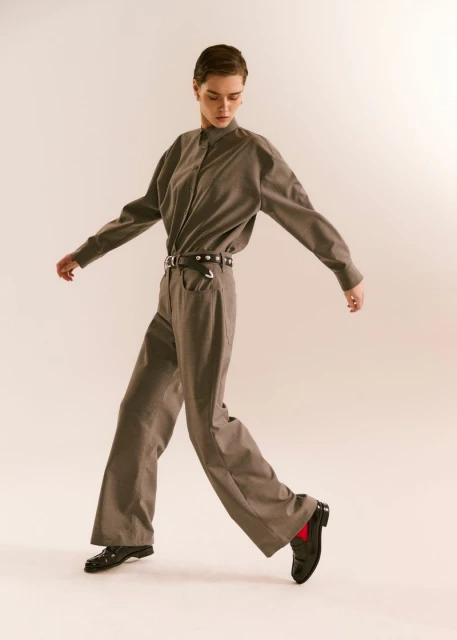 A young woman in a grey pantsuit walks forward with arms outstretched in a fashion pose against an off-white studio backdrop.