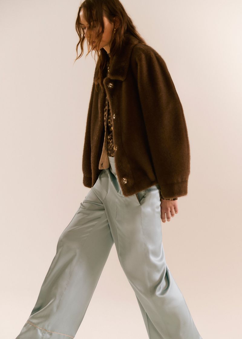 Fashionable woman in a brown jacket and light blue pants, walking in a studio setting, showcasing contemporary style.