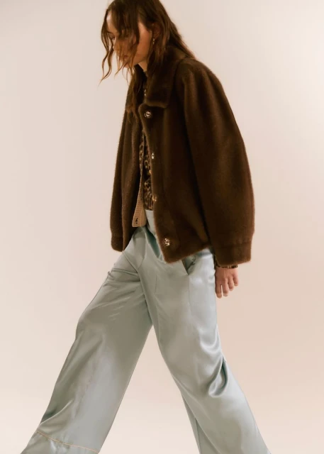 Fashionable woman in a brown jacket and light blue pants, walking in a studio setting, showcasing contemporary style.