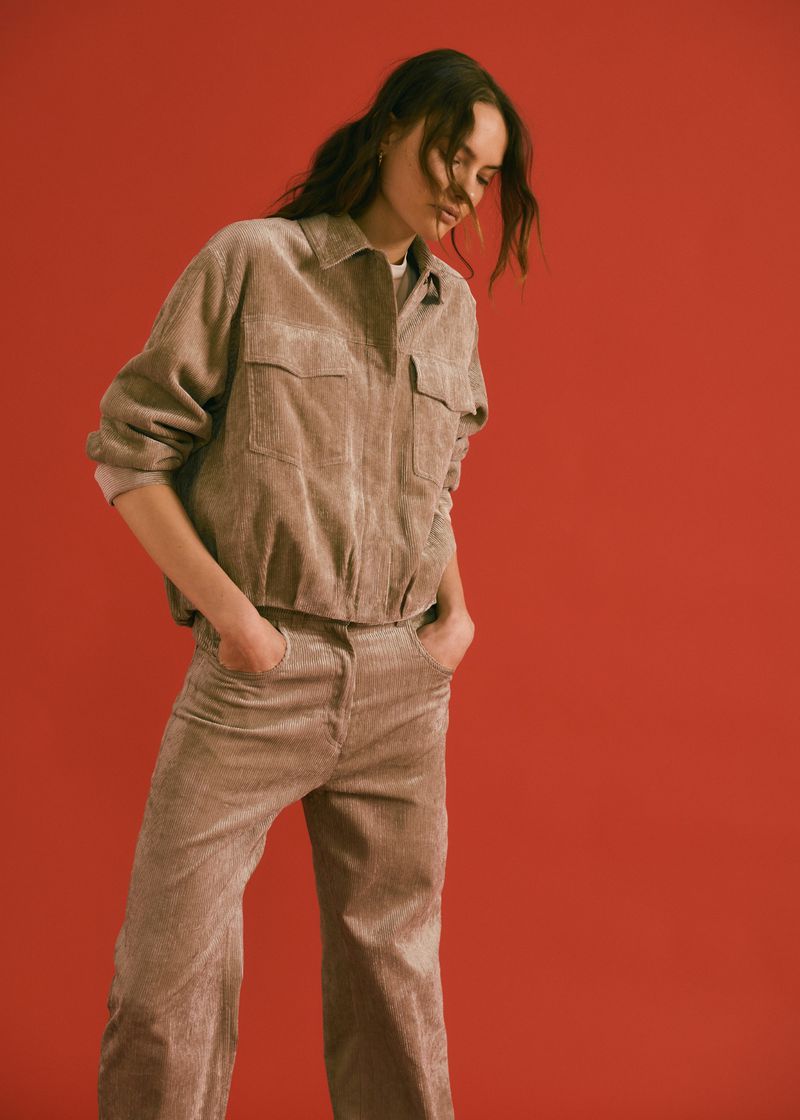 Young adult female in a corduroy jacket and trousers posing against a red backdrop in a fashion forward editorial studio shot.