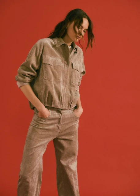 Young adult female in a corduroy jacket and trousers posing against a red backdrop in a fashion forward editorial studio shot.