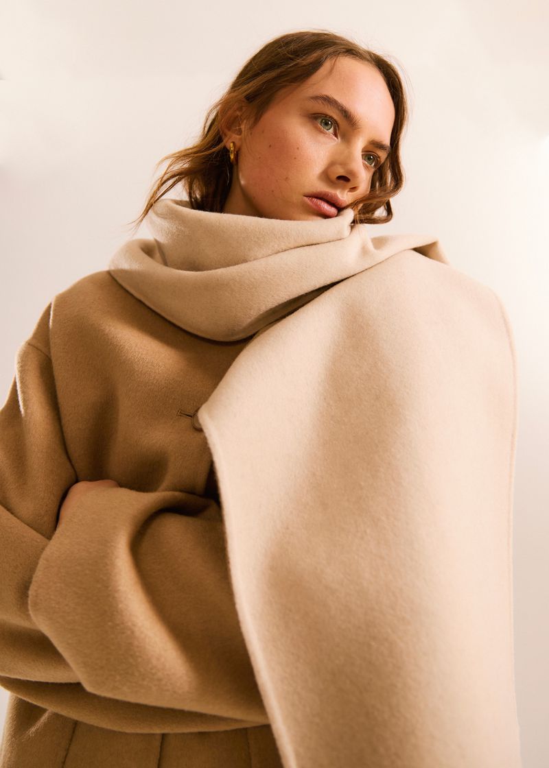 Adult woman wearing neutral-toned coat and scarf in a close-up fashion shot against a soft, diffused light background.
