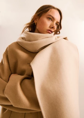 Adult woman wearing neutral-toned coat and scarf in a close-up fashion shot against a soft, diffused light background.