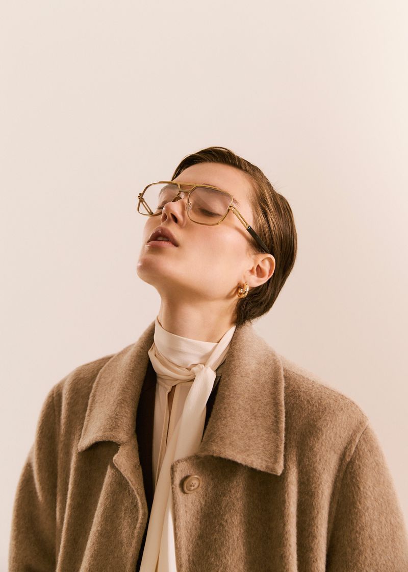 Woman with closed eyes wears glasses, a cream-colored blouse, and a wool coat in a studio setting, illuminated by soft, diffused light.