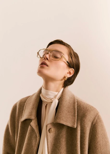 Woman with closed eyes wears glasses, a cream-colored blouse, and a wool coat in a studio setting, illuminated by soft, diffused light.