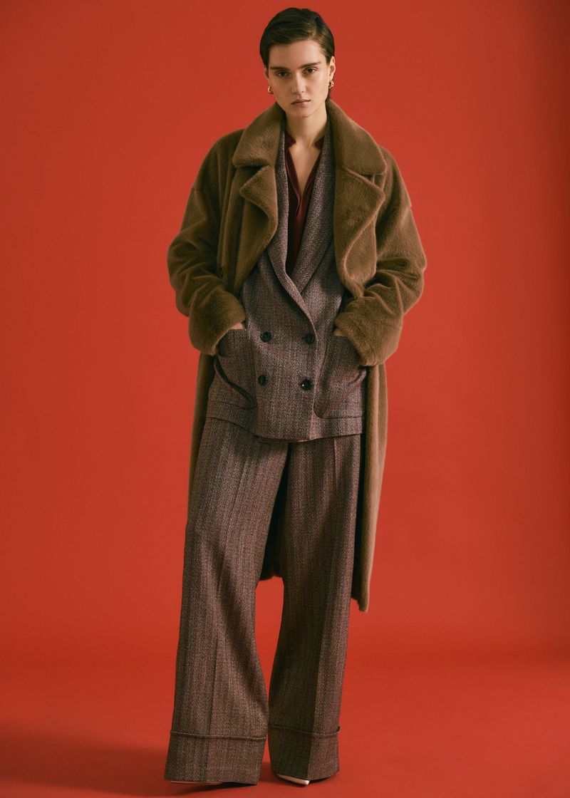 Model wearing a brown fur coat over a herringbone suit against a solid red studio backdrop