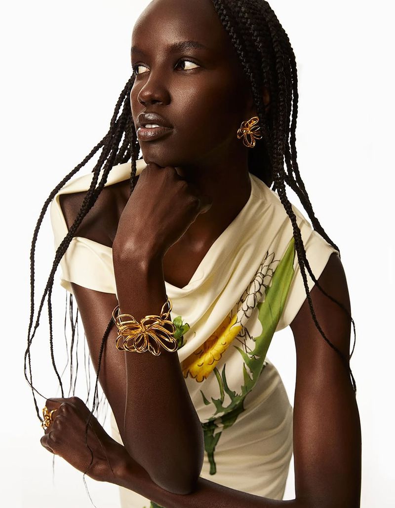 An African woman with dark skin and braided hair poses indoors, wearing a light patterned dress and gold flower jewelry against a white backdrop.