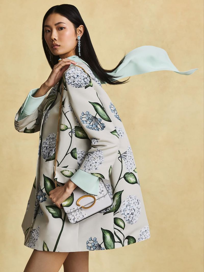 Elegant Asian woman in floral print coat holding a purse, accessorized with long earrings and a light blue scarf against yellow backdrop.