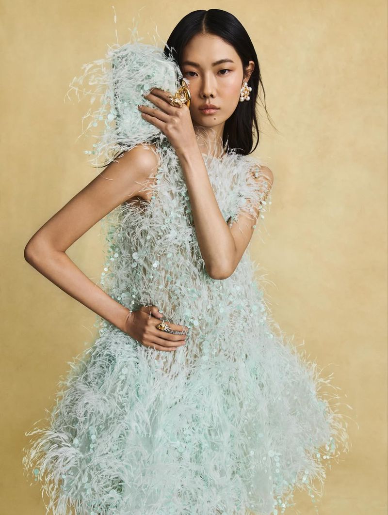 Elegant Asian woman poses in a feather dress against a gold backdrop, wearing statement earrings and a ring, showcasing high fashion.