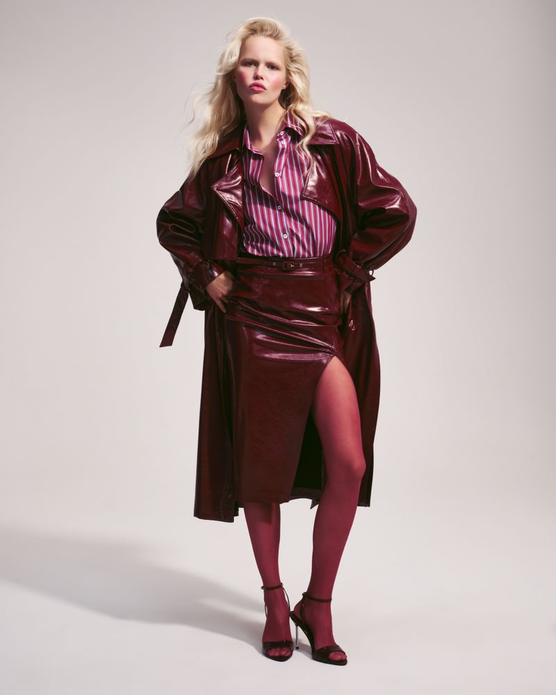 A fair-skinned woman poses in a shiny burgundy trench coat and skirt with a vertical striped shirt, hose and high heels against a pale background.