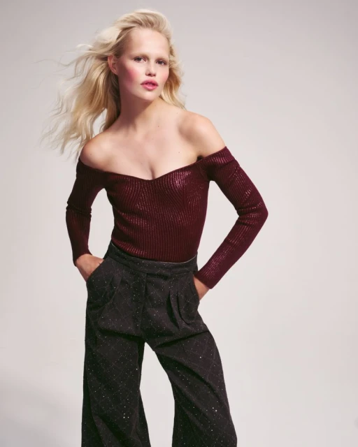 A fair-skinned fashion model with blonde hair poses in a studio wearing a maroon off-the-shoulder top and wide-leg patterned pants.