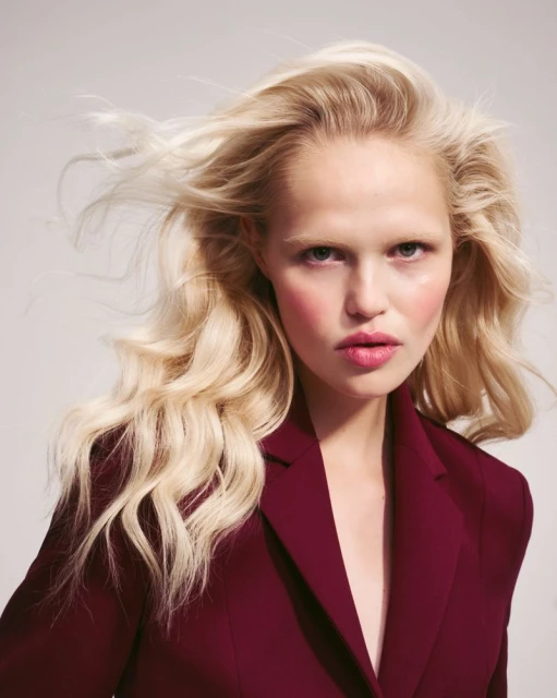Close-up portrait of a fair-skinned adult female with flowing blonde hair and wearing a burgundy blazer in a studio setting.