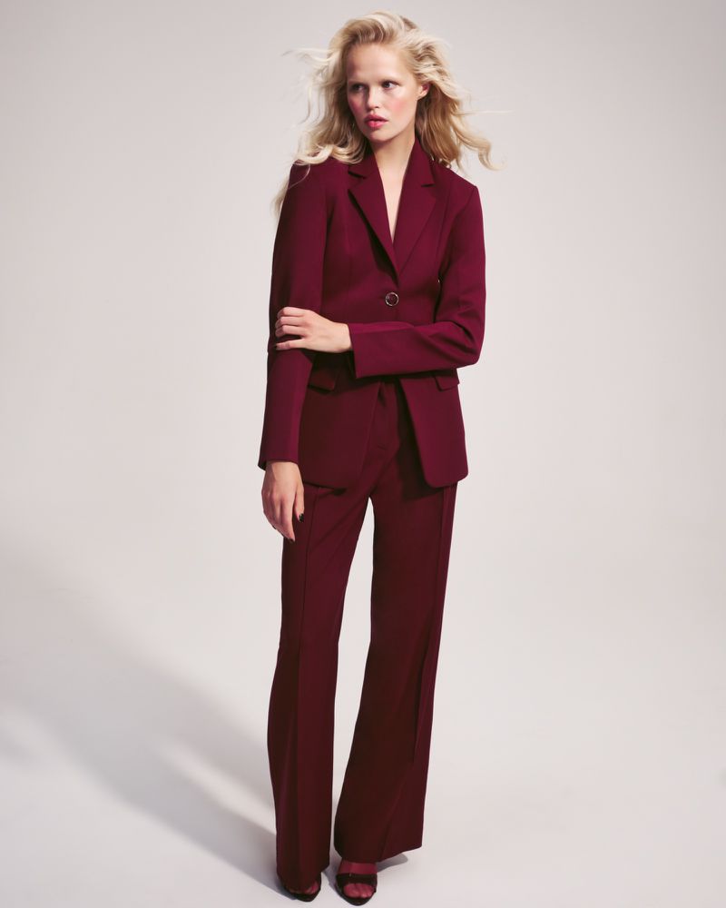 Model wearing a structured burgundy blazer and matching trousers against a pale neutral studio backdrop.