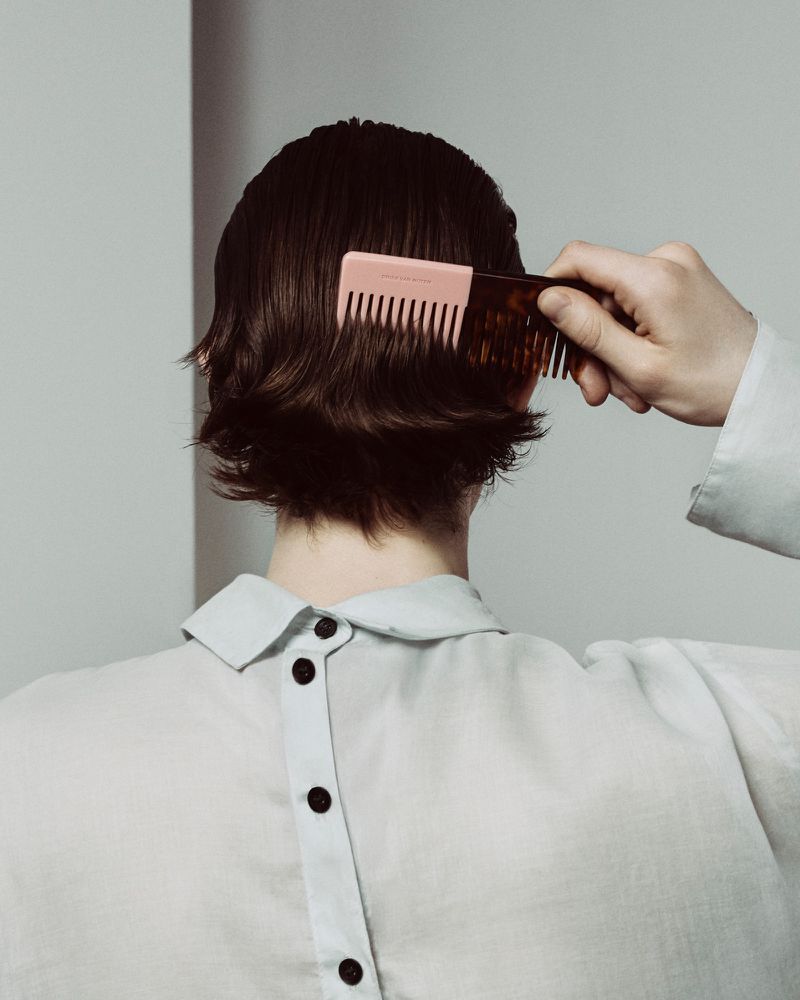 Person with short dark hair, back to camera, wearing a collared shirt and combing hair with a two-toned comb on light grey.