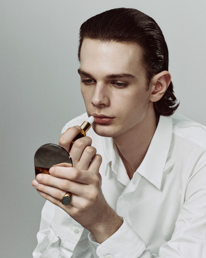 Close-up portrait of a man applying lipstick with a compact mirror wearing a white collared shirt against a neutral background.