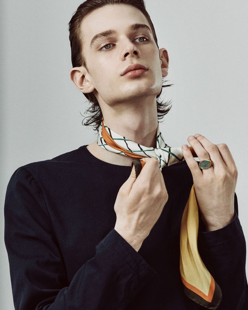 Indoor portrait of a light-skinned adult male tying a patterned neck scarf, wearing a black long-sleeve shirt, against a gray backdrop.