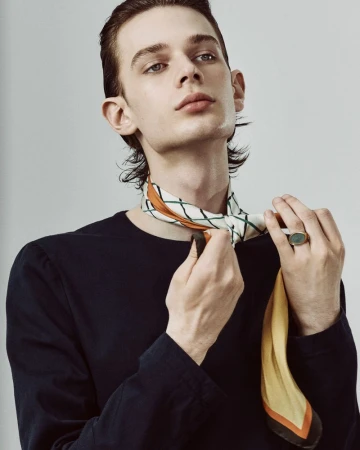 Indoor portrait of a light-skinned adult male tying a patterned neck scarf, wearing a black long-sleeve shirt, against a gray backdrop.