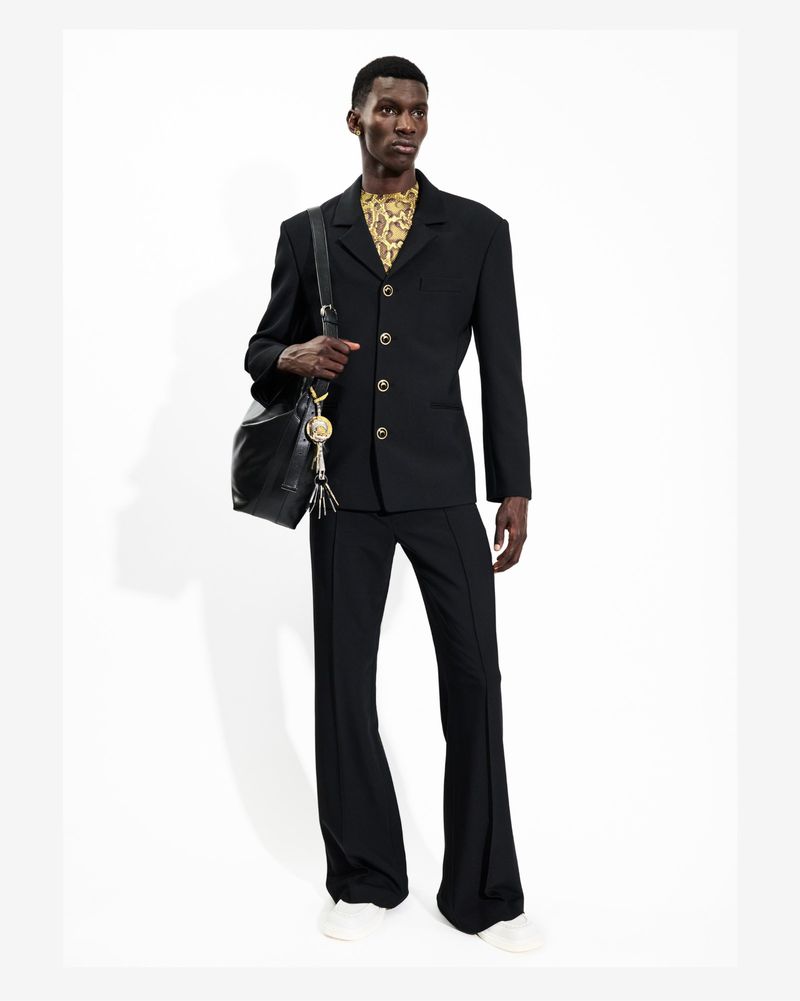 A stylish man in a black suit and snakeskin-patterned shirt poses in a photo studio, carrying a black leather bucket bag, showcasing fashion.