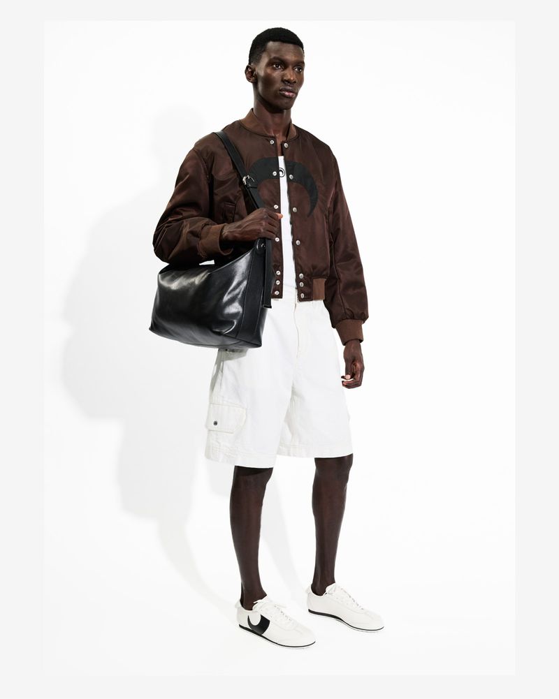A Black male model poses in a studio wearing a brown bomber jacket, white shorts, and sneakers, with a black messenger bag over his shoulder.