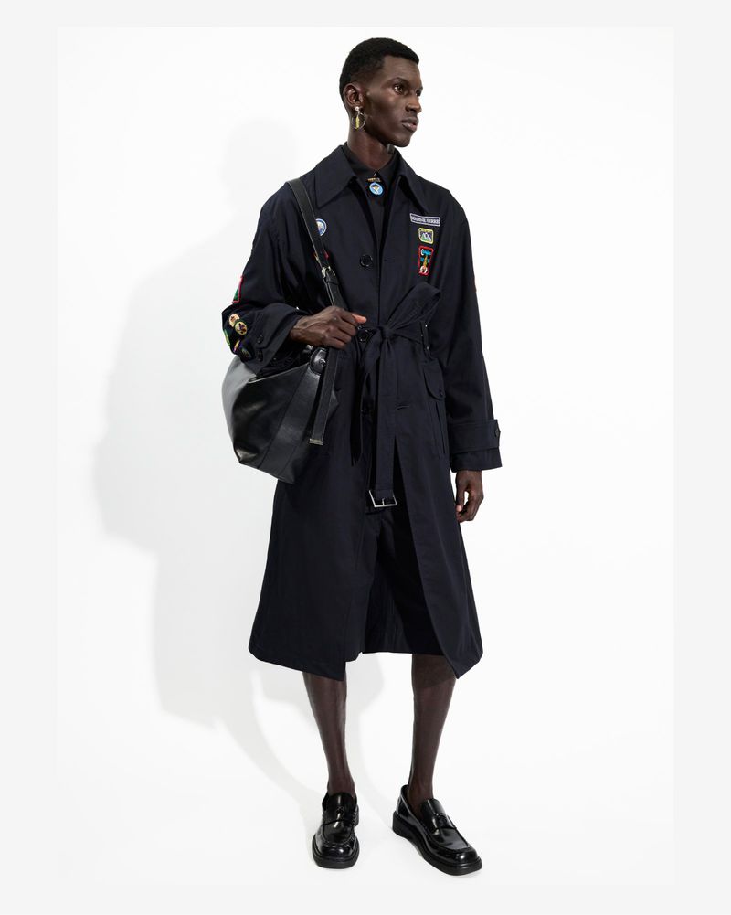 A man in a dark trench coat and loafers stands in a photo studio wearing a black shoulder bag against a plain white background.