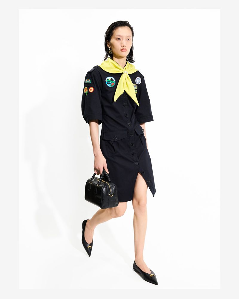 Woman in navy shirt dress with a yellow neckerchief, black handbag, and black flats on a white backdrop in a studio shot.