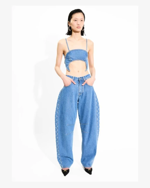 An Asian woman poses in a light blue denim crop top and oversized jeans with lace-up details, accessorized with hoop earrings and black heels.