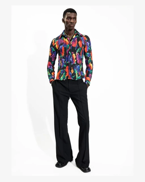 Full-length shot of a Black male fashion model in a retro floral shirt and black bell-bottom pants, posing in a white photo studio.