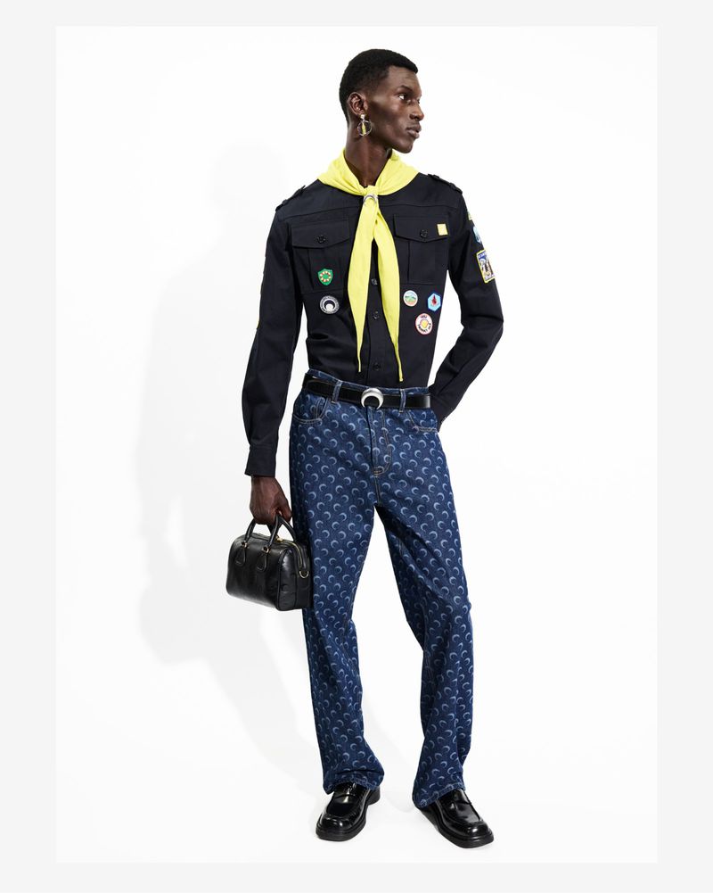 Dark-skinned male model in a black shirt, patterned blue pants, and black shoes, holding a small black bag against a white background.
