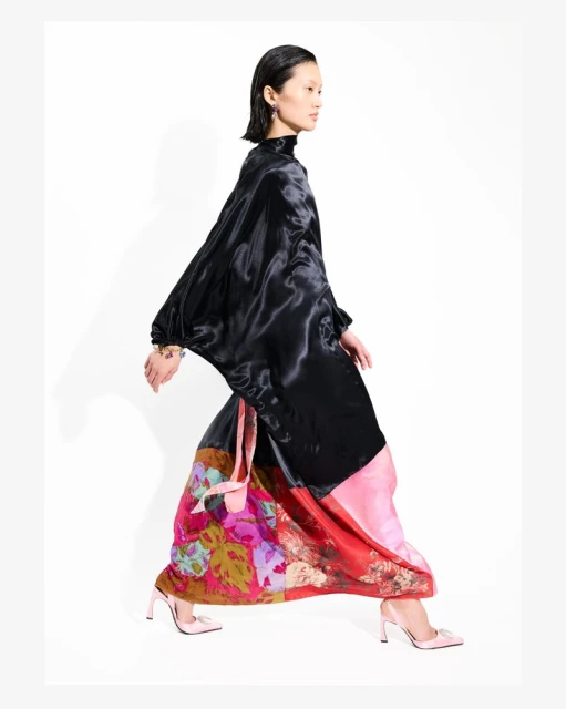 Asian woman in shiny black top and colorful patterned skirt, wearing pink heels, poses in a full shot in a brightly lit photo studio.